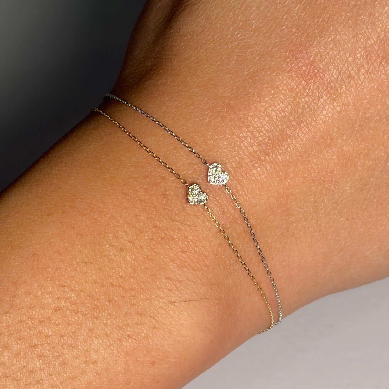 Close-up of a wrist wearing two delicate bracelets with heart-shaped charms on a neutral background.