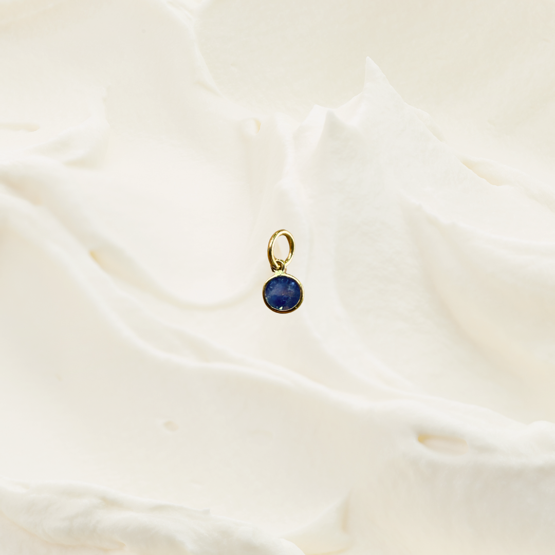 A small gold pendant with a round dark blue gemstone, resting on a softly textured, cream-colored fabric background.