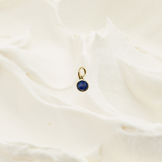A small gold pendant with a round dark blue gemstone, resting on a softly textured, cream-colored fabric background.