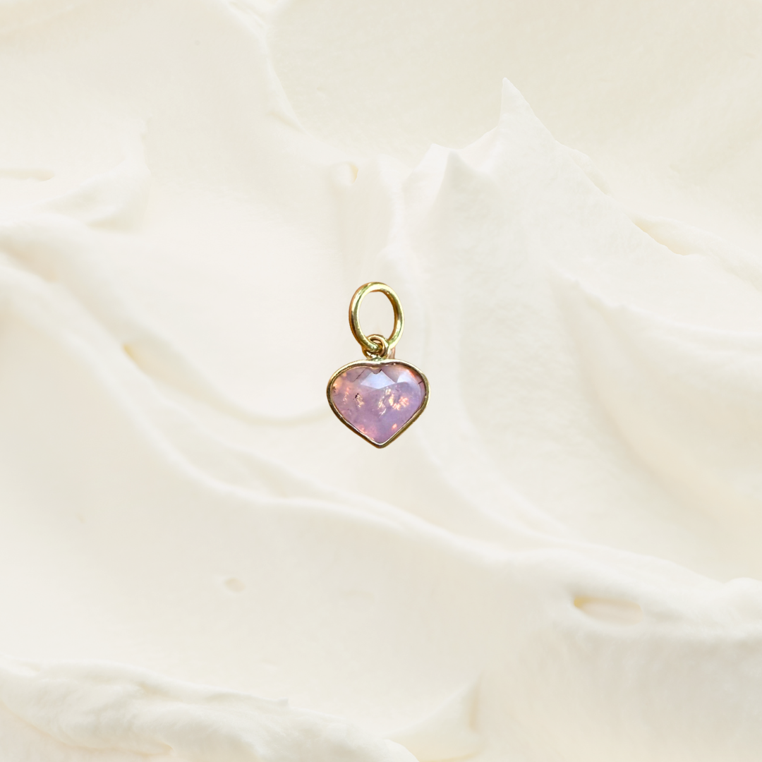A small gold heart-shaped pendant with a pink iridescent opal center, resting on a soft, draped cream fabric background.