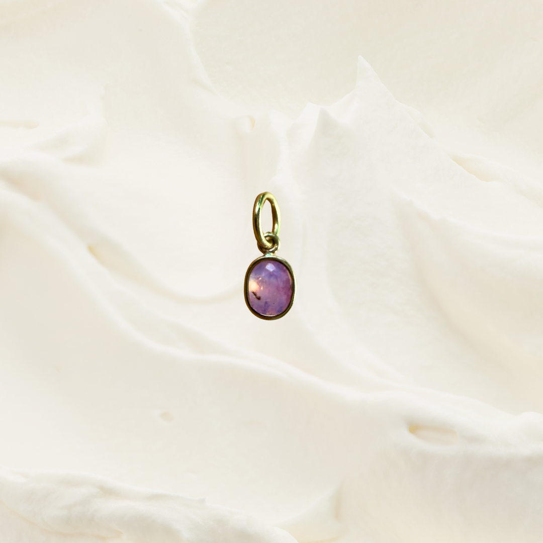 A small oval pendant with a purple stone and gold-tone bezel, resting on a softly draped white fabric background.