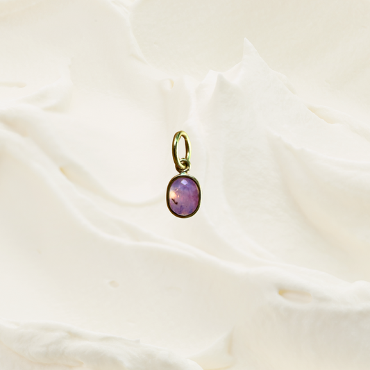 A small oval pendant with a purple stone and gold-tone bezel, resting on a softly draped white fabric background.