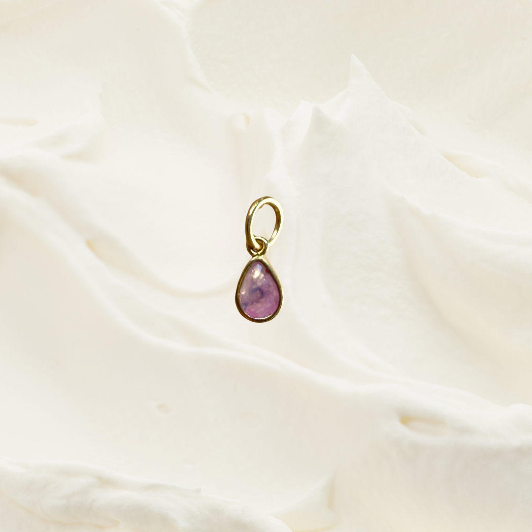 A small gold teardrop pendant with a pinkish-purple stone, resting on a soft, textured white fabric background.