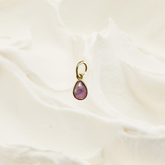A small gold teardrop pendant with a pinkish-purple stone, resting on a soft, textured white fabric background.