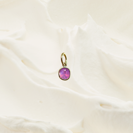 A small gold pendant with a round pink gemstone, resting on a textured, softly draped off-white fabric background.