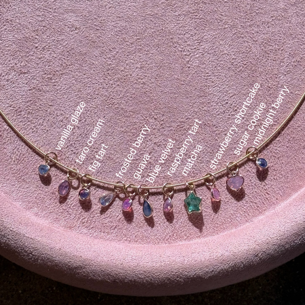 A gold-tone necklace with multiple small teardrop-shaped gemstone charms in assorted colors, displayed on a pink velvet jewelry stand. The necklace has white text labels identifying each gemstone type, including pomegranate, blue cream, fig leaf, toasted berry, ice, raspberry ketone, mint, strawberry shortcake, and chocolate chip.